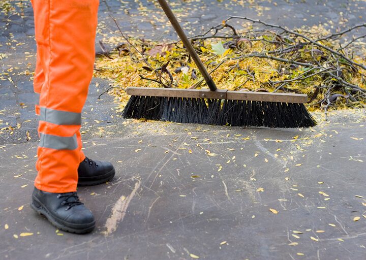 A person wearing bright orange work pants sweeping leaves with a large push broom